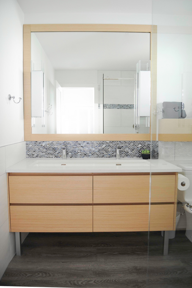 Bathroom vanity beneath small grey tile back splash and large wood frame mirror.