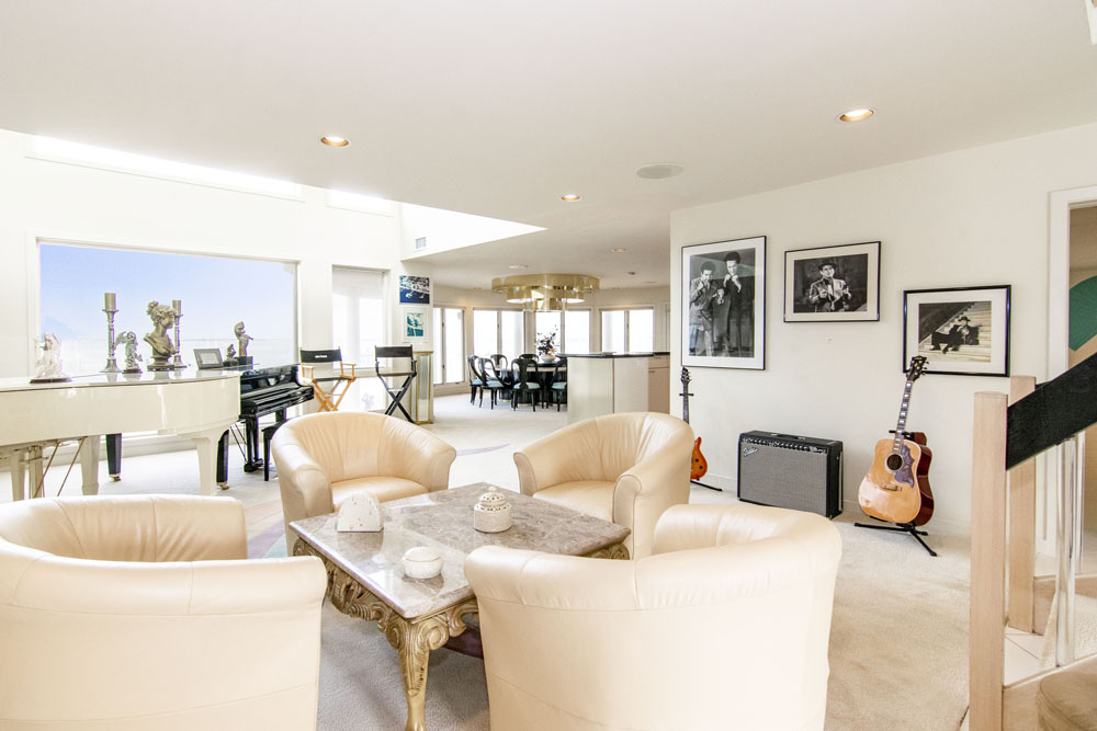 A white open-concept living room and dining area with a grand piano and floor-to-ceiling windows overlooking the water