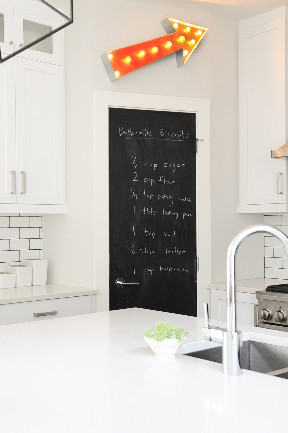 A monochromatic white kitchen with a basement door chalkboard and LED arrow sign