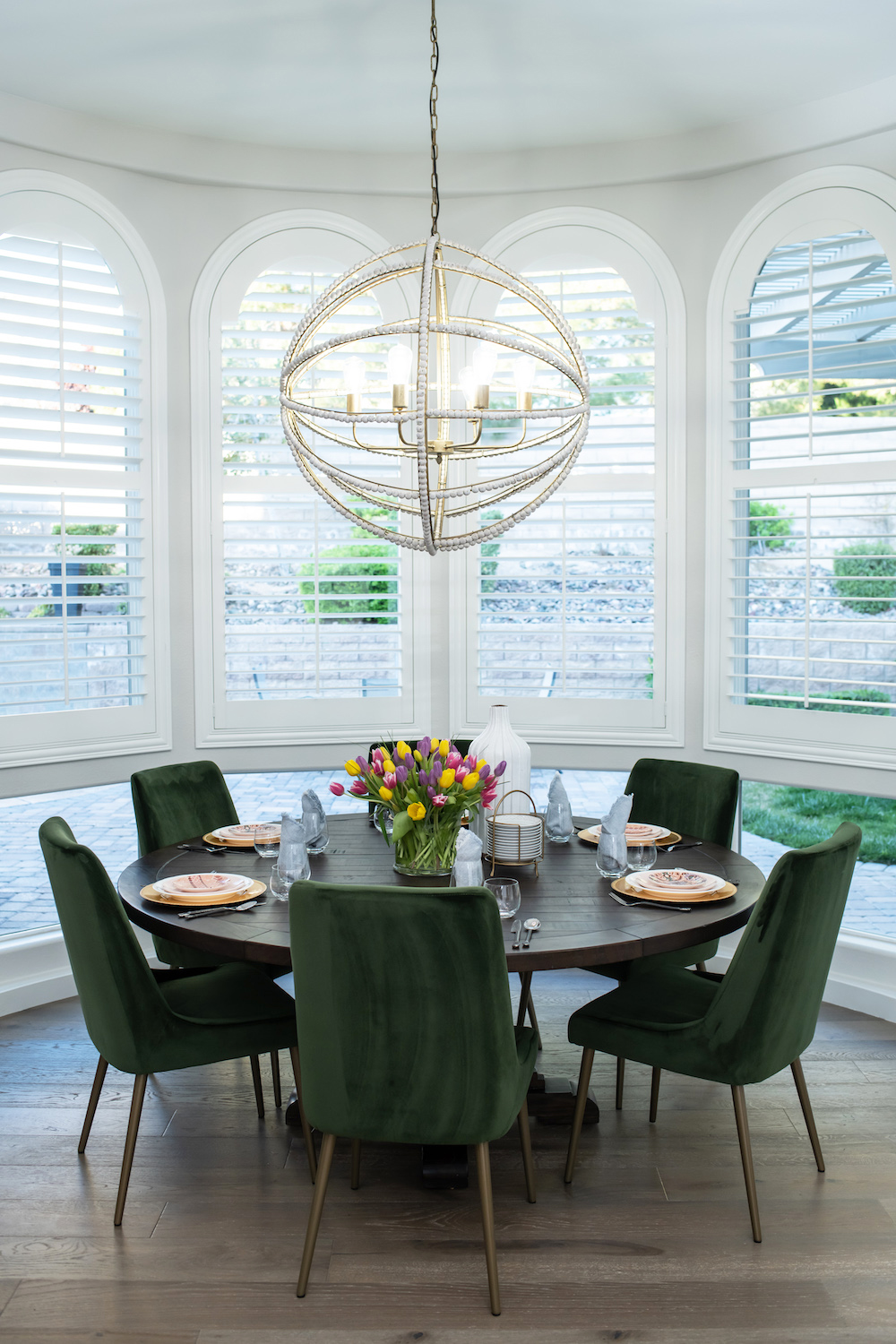 A dining room next to bay windows with a large circular table, offbeat chandelier and stunning plush green chairs