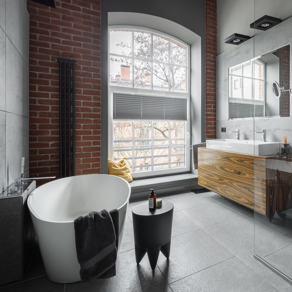 industrial-style bathroom with oval bathtub, grey tiled floor, brick wall and big window