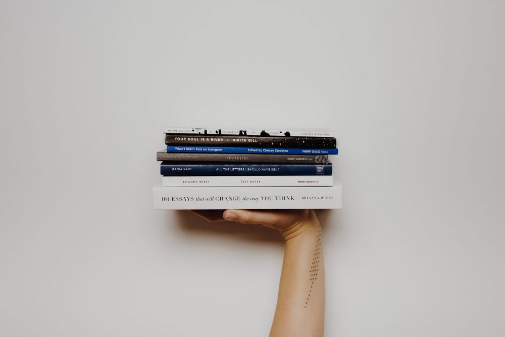 A woman's tattooed arm holds up a stack of books against a white background