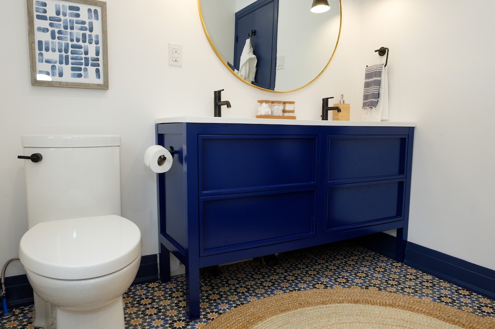 white bathroom with round mirror, blue vanity and small glass storage containers