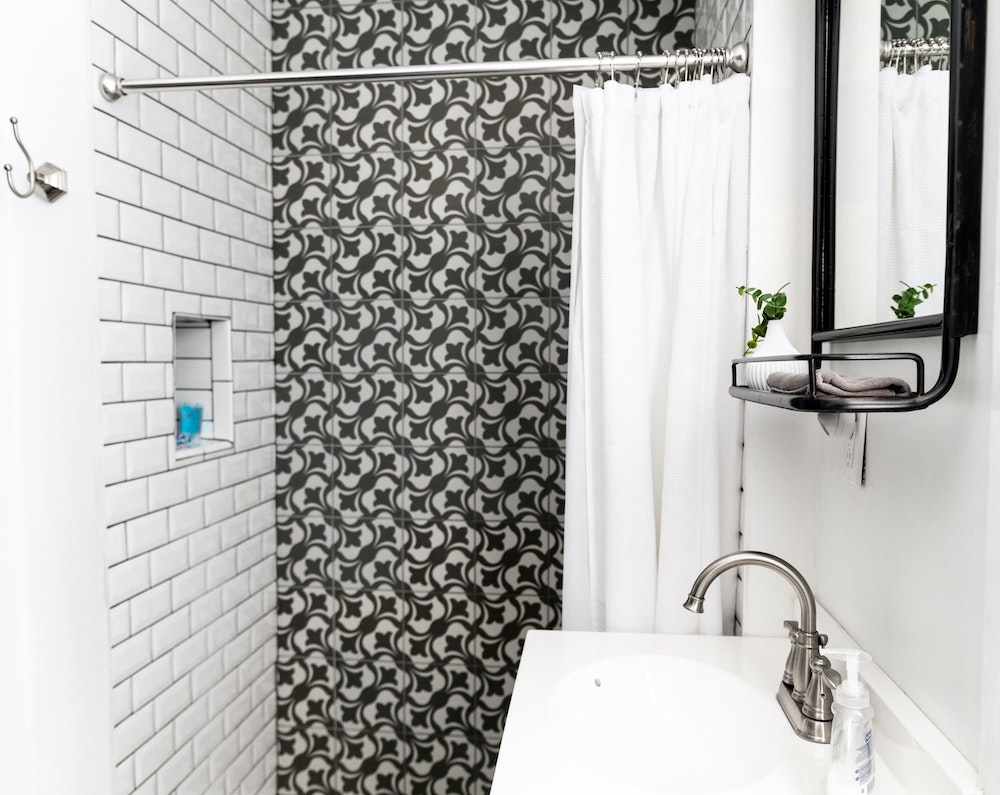 small bathroom with patterned-tile shower and small white sink with black mirror with shelf