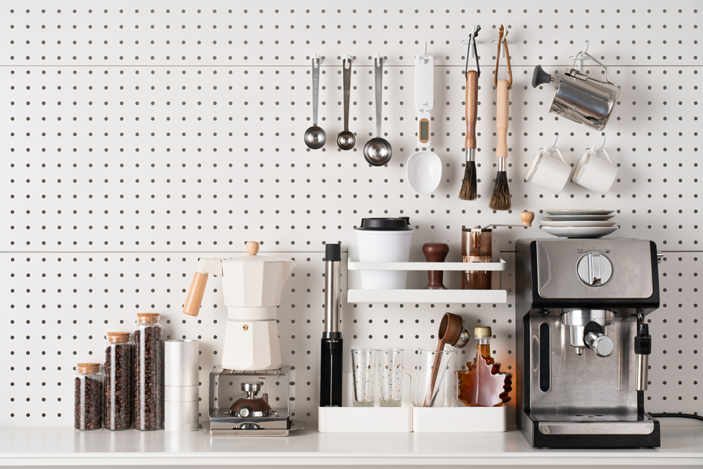 A large white pegboard against a wall in front of a coffee maker, espresso machine and other various appliances