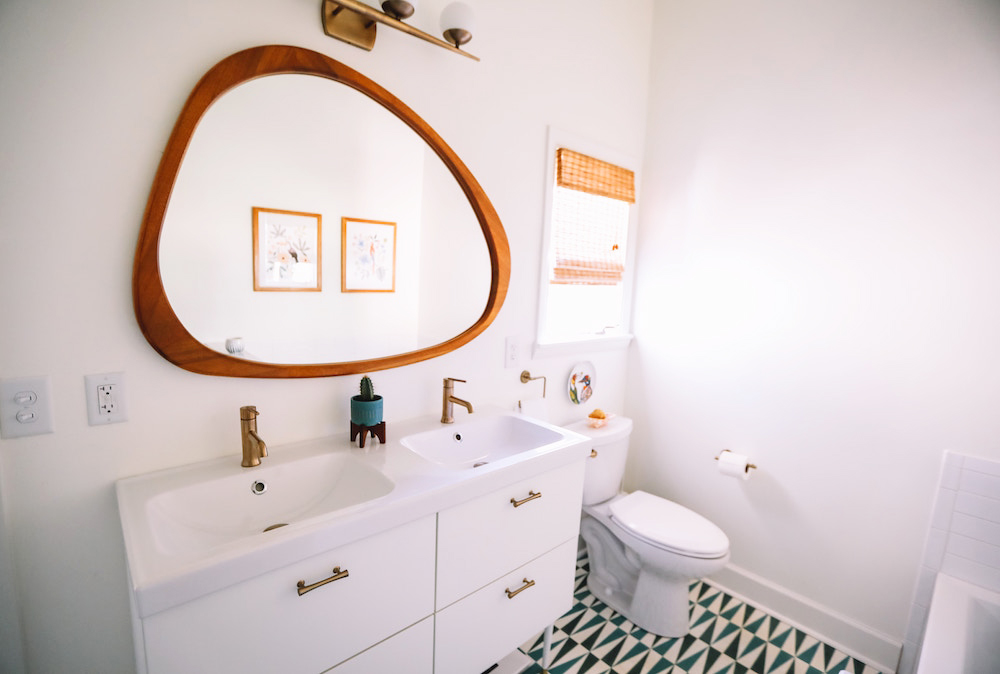 A small white bathroom with patterned tile floors, abstract mirror above the sink and minimal decor