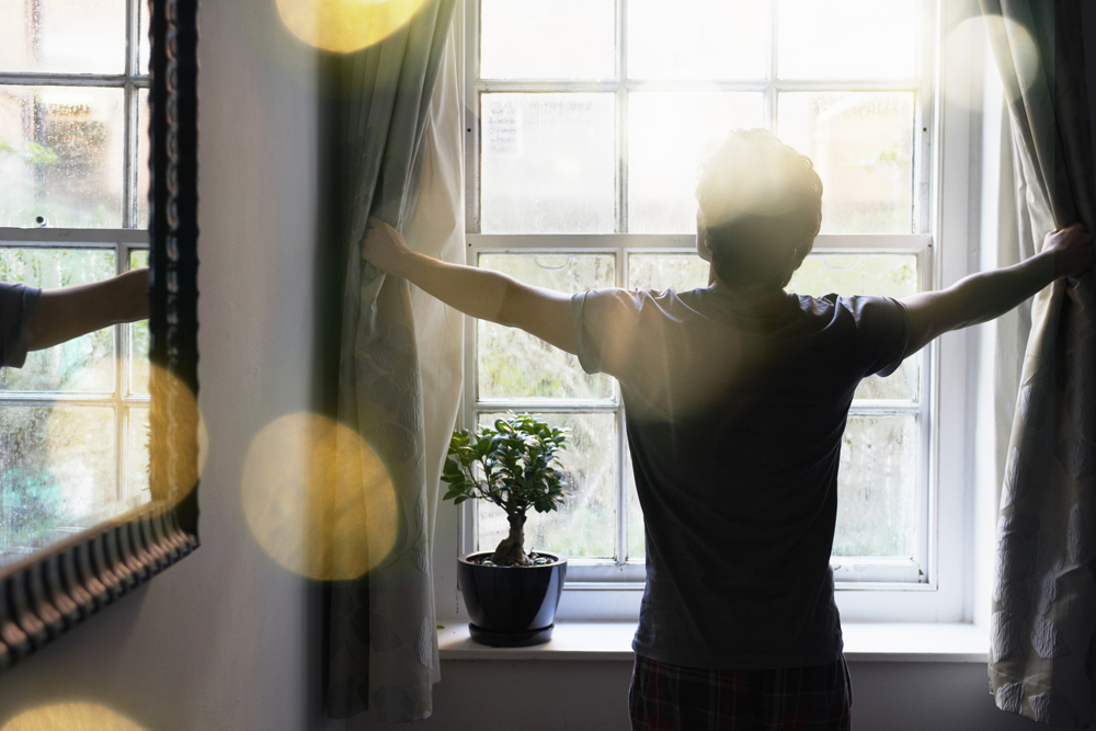 A man opens his bedroom curtains to let in the morning sunshine