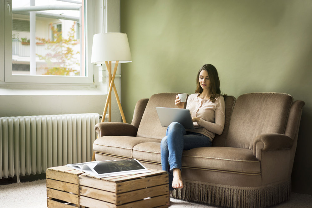Young woman sitting on couch with cup of coffee using laptop with a wooden crate as her coffee table