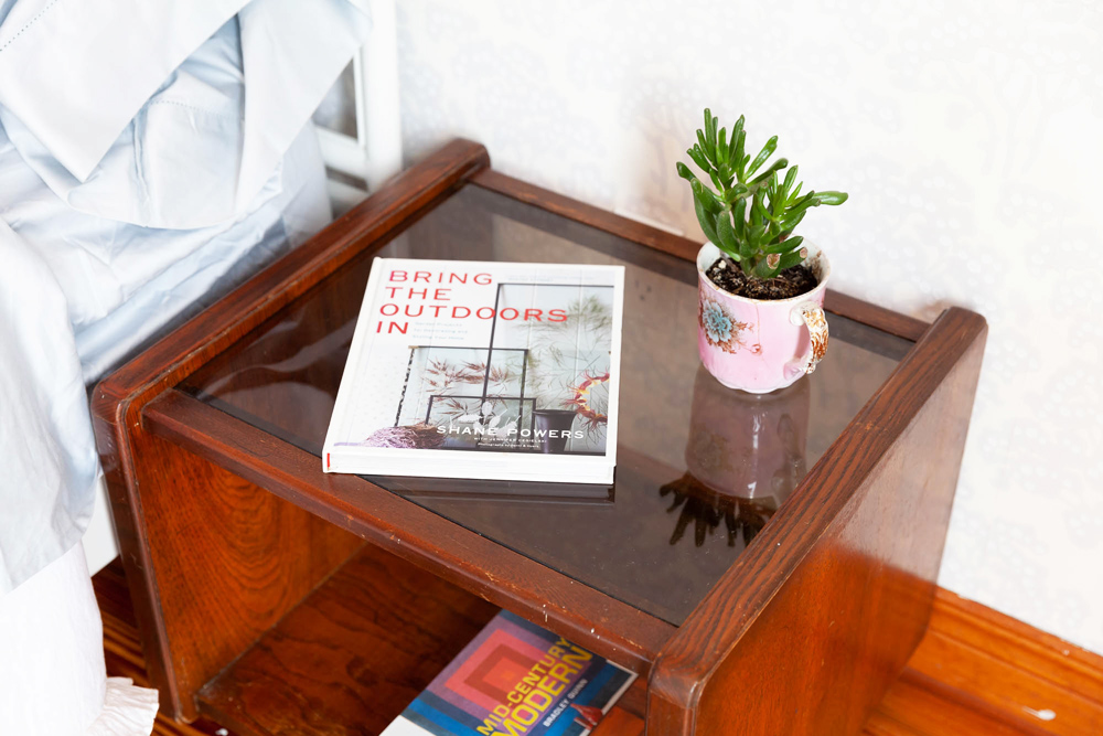 A thrifted wood bedside table with books and small plants on its glass surface