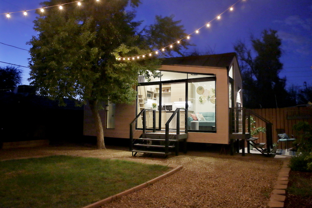 tiny house with large glass windows, sits in the backyard at night, surrounded by trees and party lights