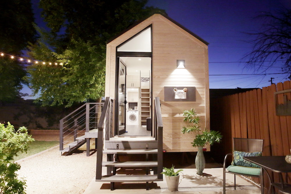tiny house with large glass windows, sits in the backyard at night, surrounded by trees and party lights