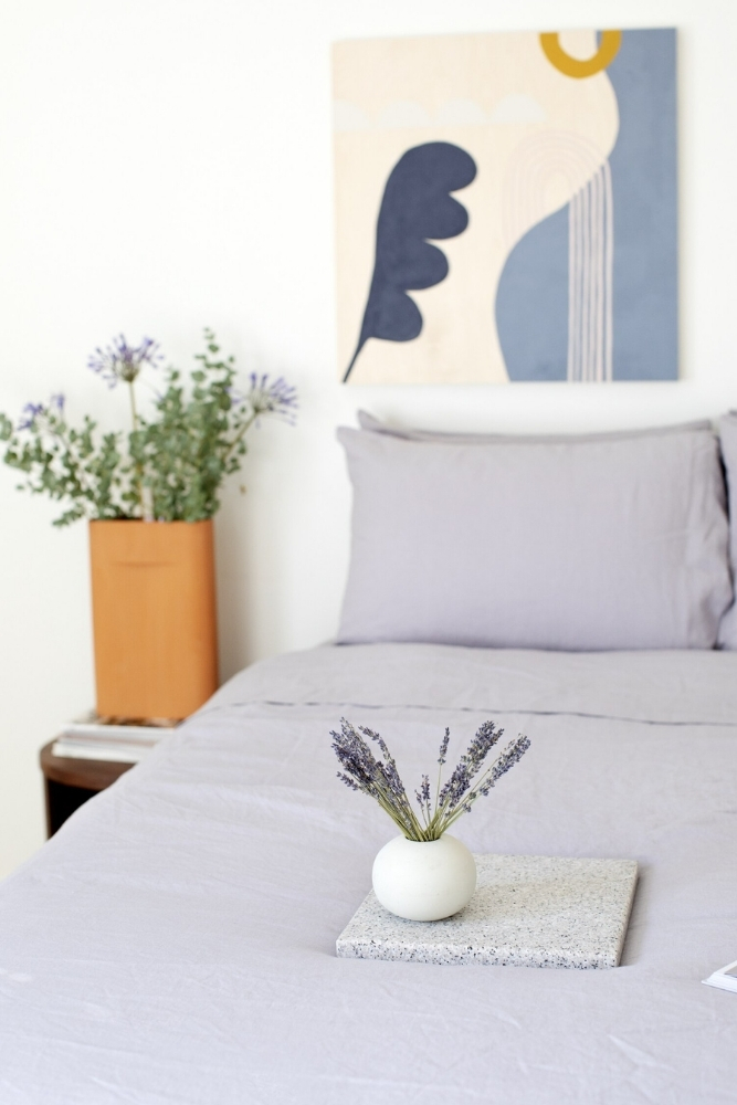 A bed with linen bed sheets in lavender. Also, a vase of lavender flowers on the bed.