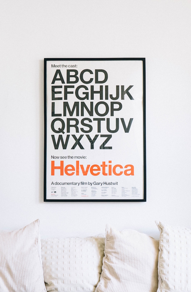 white wall with black-framed Helvetica movie poster and white couch with pillows