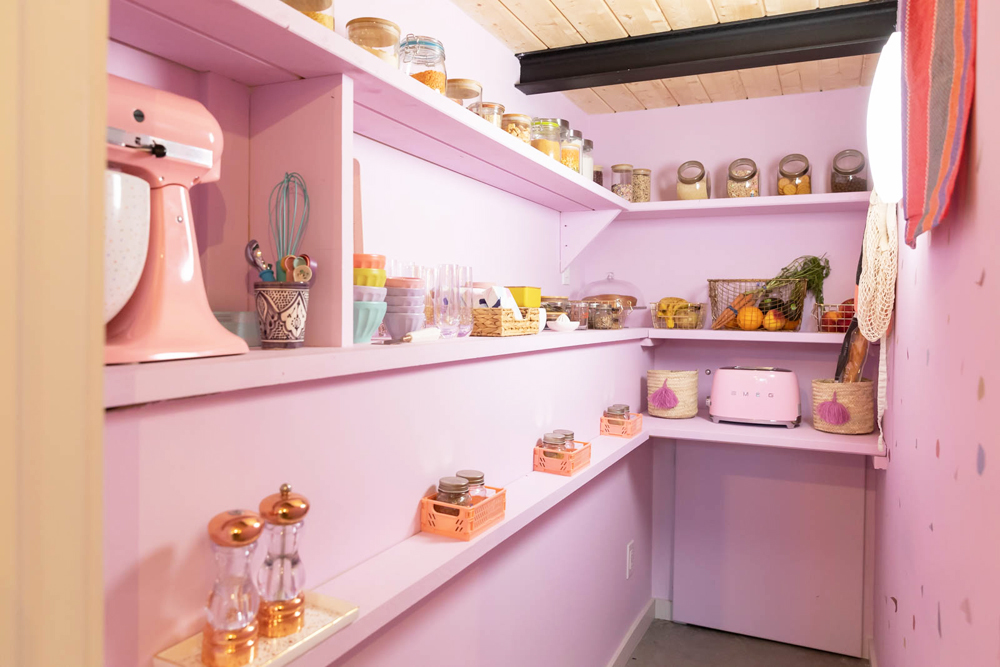 A narrow pastel pink walk-in pantry with matching appliances and glass jars