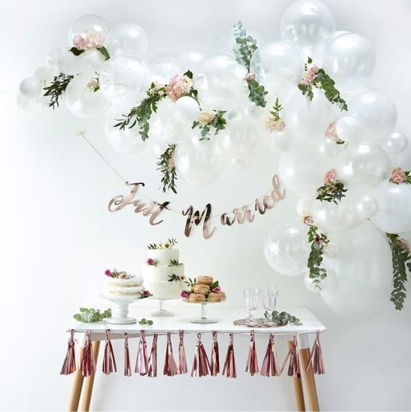 Clear balloon arch with green foliage above a table with wedding desserts