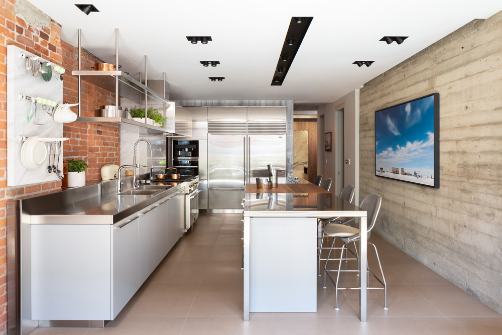 view to kitchen, exposed brick wall on left, cement wall with painting on right, island, stainless steel fridges in back