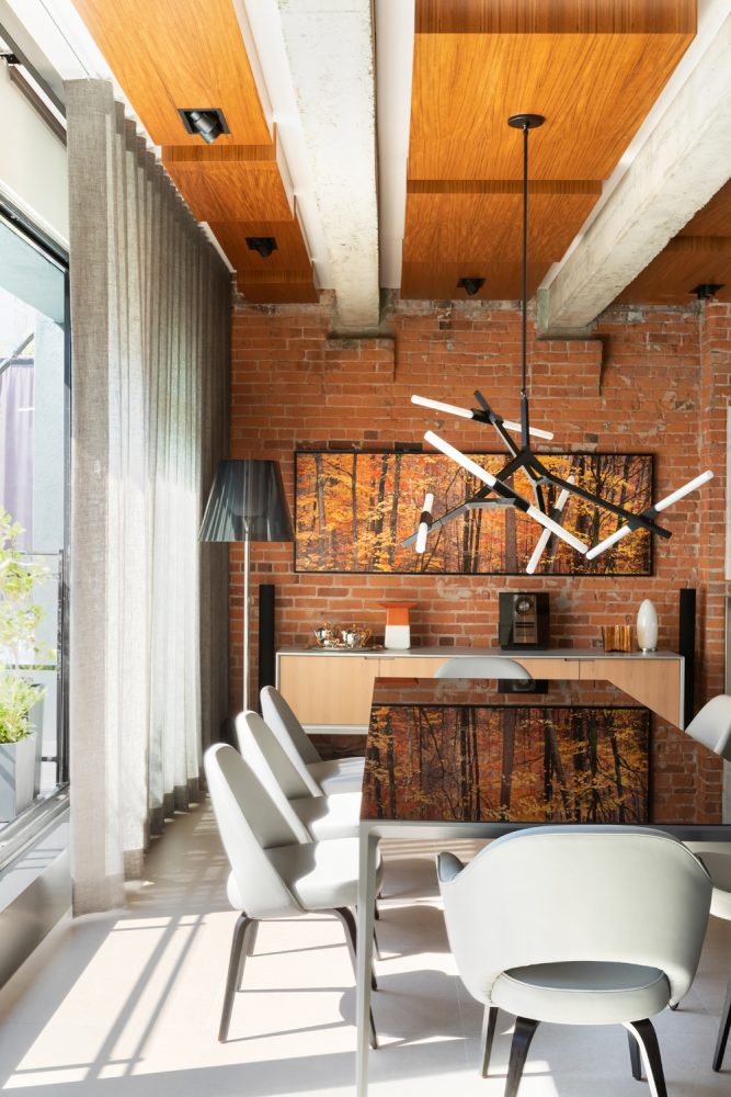 loft dining room with exposed brick wall, table top reflecting tree painting and sculpted grey dining chairs