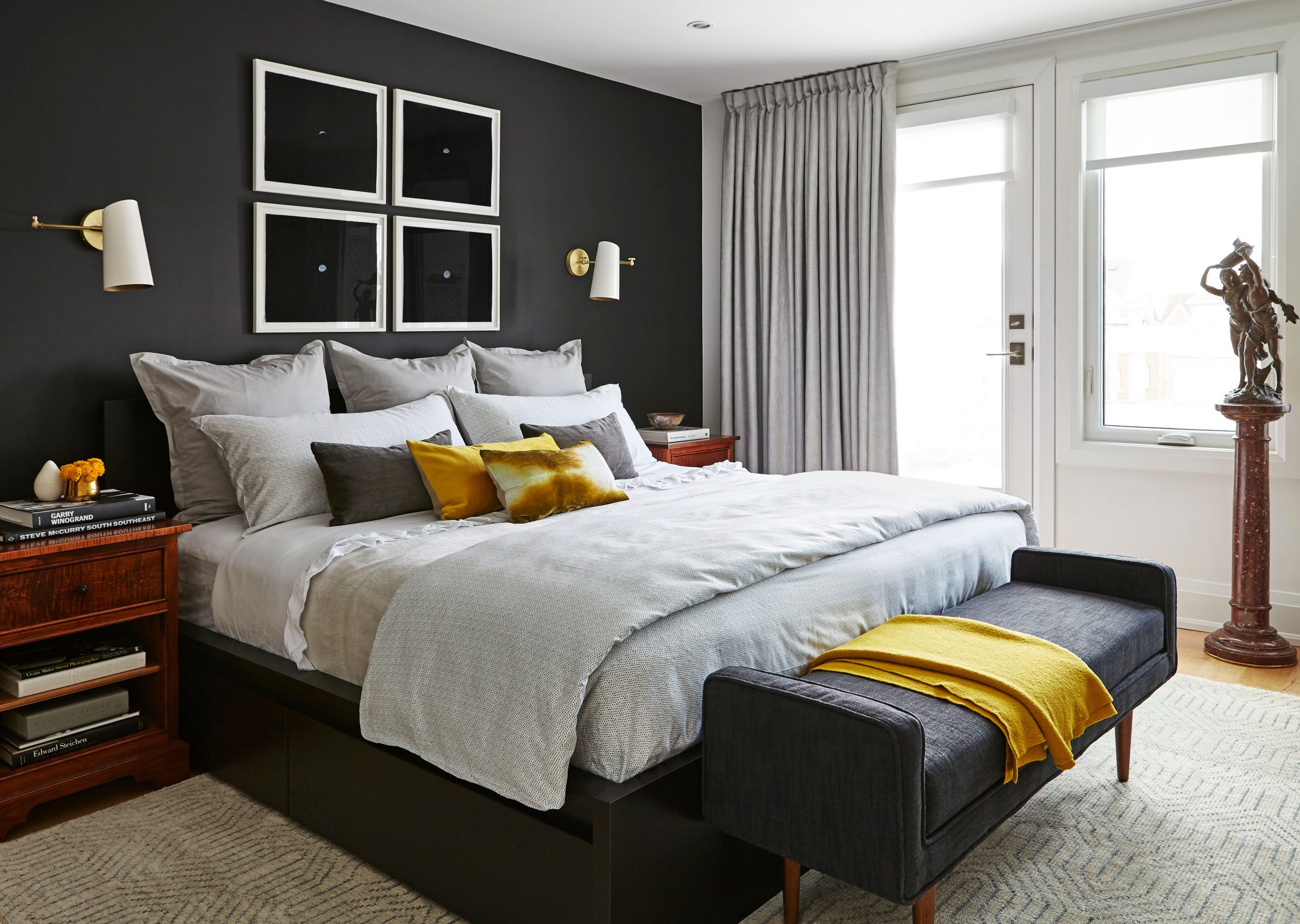 bedroom with black bed, black focal wall, grey bedding with mustard accents, statue on marble pedestal by window