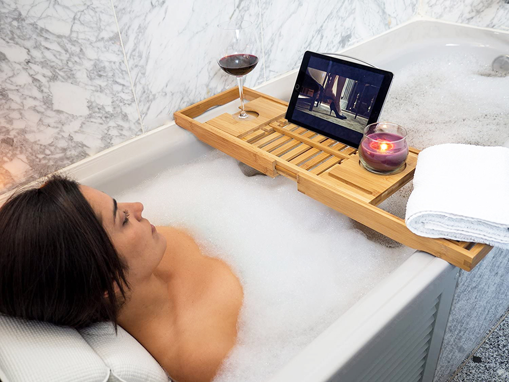 A woman leans back and soaks in the tub while drinking red wine and watching a TV show on an iPad while balancing items on a wooden bathtub caddy