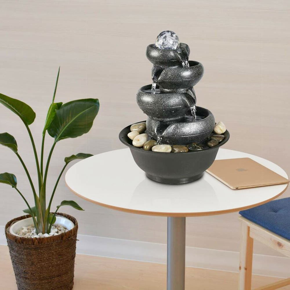A grey three-tiered cascading fountain on a side table near a closed laptop and potted plants