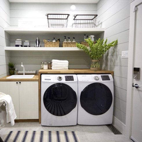 Pretty white laundry room with grey tile floors, a black and white striped rug, a white washing machine and dryer, wooden folding counter and open shelves with laundry soap and accessories as featured on HGTV Canada