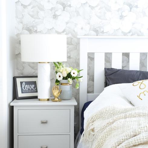A bedroom with floral wallpaper, a white bed and gray night stand.