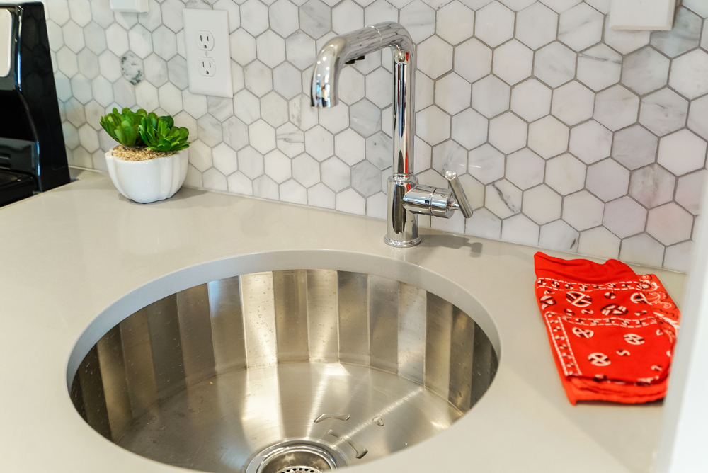 Sleek and small circular stainless steel sink with small hexagon tile backsplash and grey quartz countertop as seen on Masters of Flip on HGTV Canada