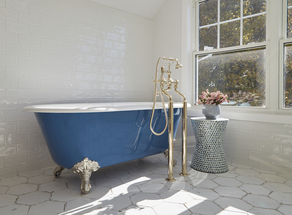 A blue clawfoot bathtub with antique taps featuring English gold stands in an all-white room with modern brick tile on the wall, and a large window as featured HGTV Canada