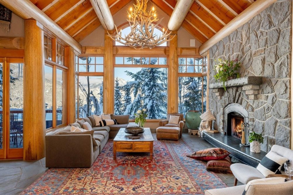 A spacious living room with a red rug, stone fireplace, massive windows and wood detailing.
