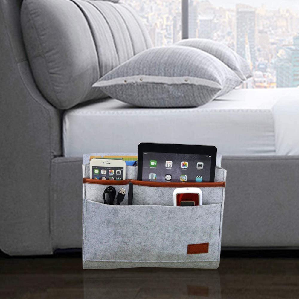 Grey Markkeer Bedside Caddy hanging from a grey upholstered bed with 4 pockets filled with iPhone, tablet, cords, and magazines