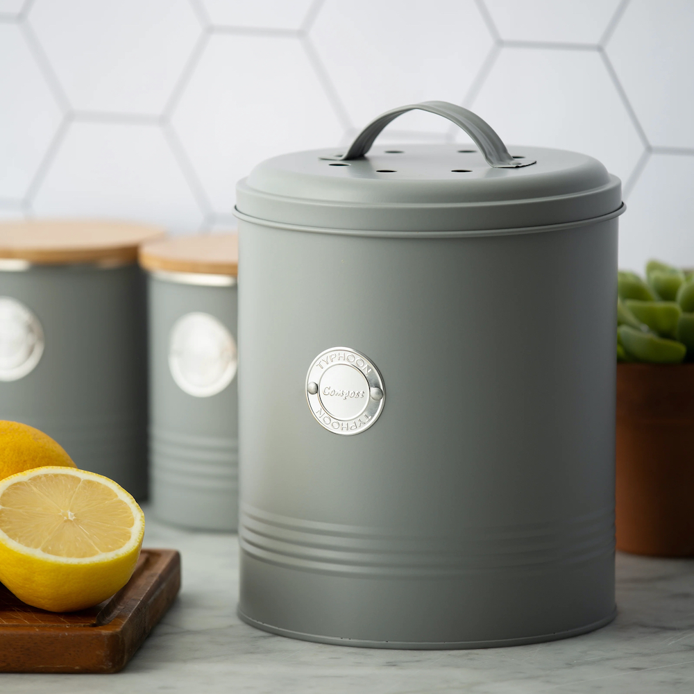 The Typhoon Living Compost Caddy sits alongside matching storage canisters on a grey marble countertop against a white hexagonal tile backsplash.