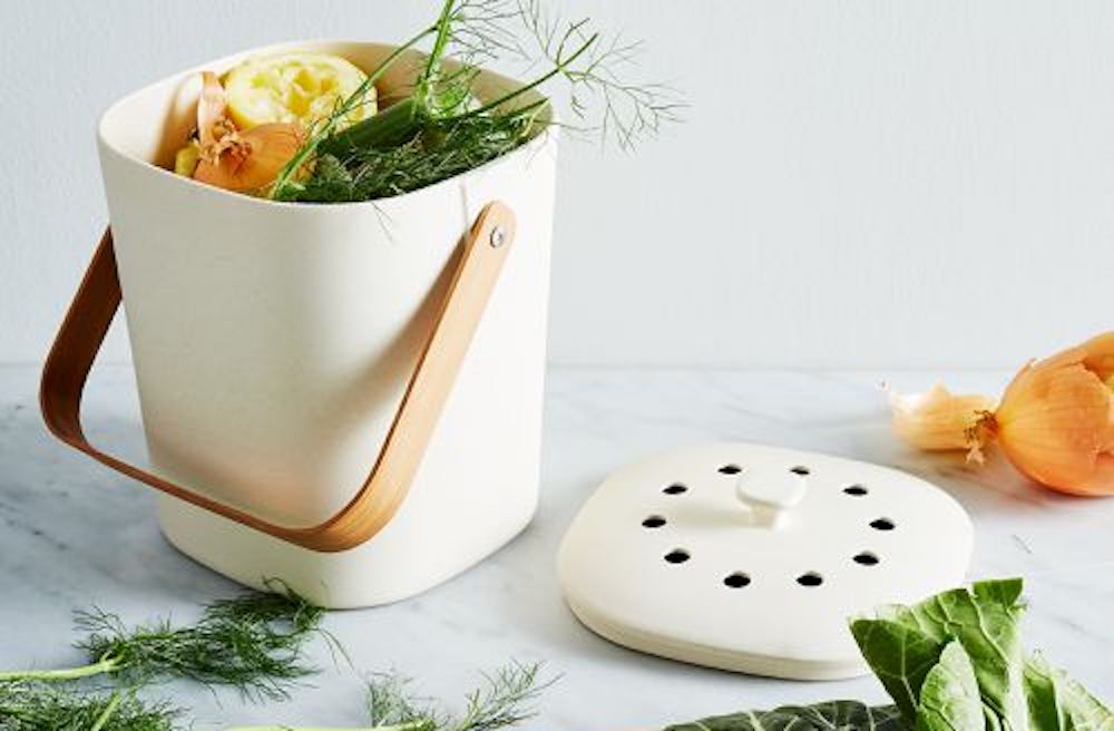 Bamboozle Countertop Compost Bin made from biodegradable bamboo fibre sits on a grey and white marble countertop and is filled and surrounded by various food scraps.