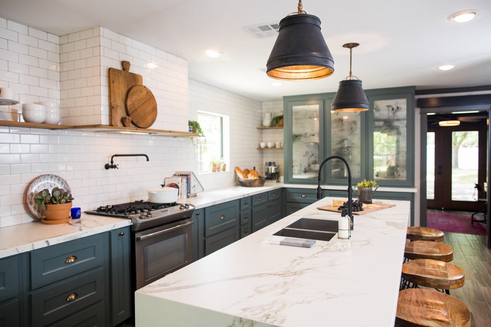 A beautiful large kitchen with huge centre island, granite countertops, white subway tile backsplash, two large black pendant lights, and cyan painted cabinets with gold hardware.