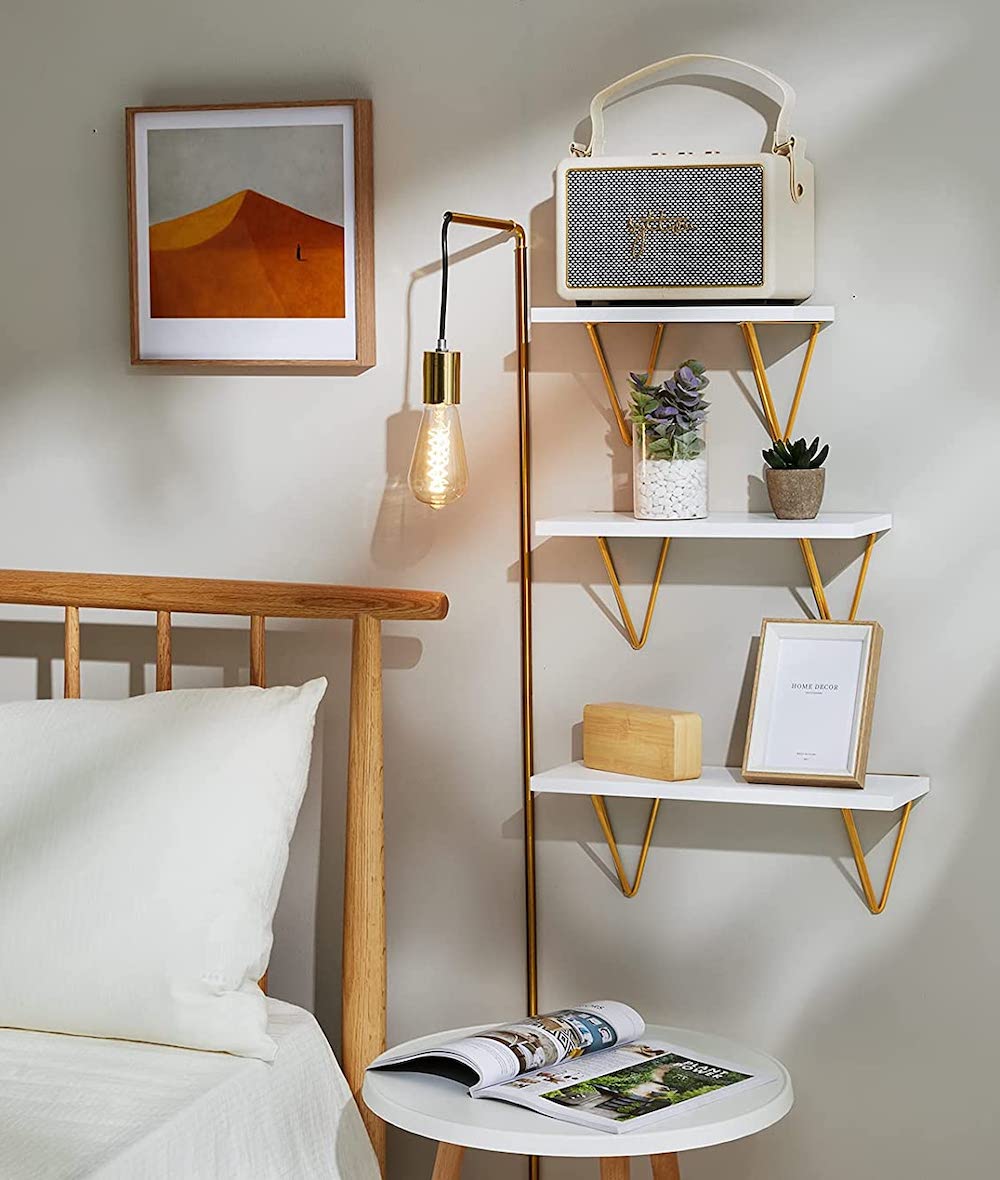 Amada Homefurnishing white floating shelves with triangle metal brackets and a gold finish sit on a wall above a bedside table beside a wood framed bed and a gold wall sconce with a filament lightbulb