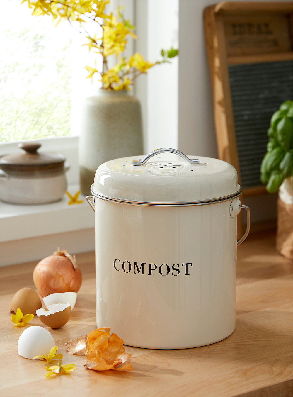 A chic, eco-friendly white enameled compost bin with ‘COMPOST’ written on it sitting on a wood countertop surrounded by food scraps