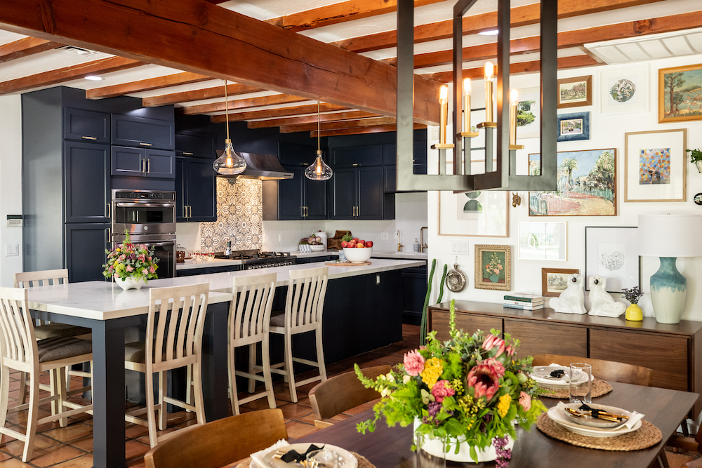 A charming kitchen with dark blue cabinets and a long eat-at island with white countertops, and a dining room with a wooden table and chairs and modernist light fixture above