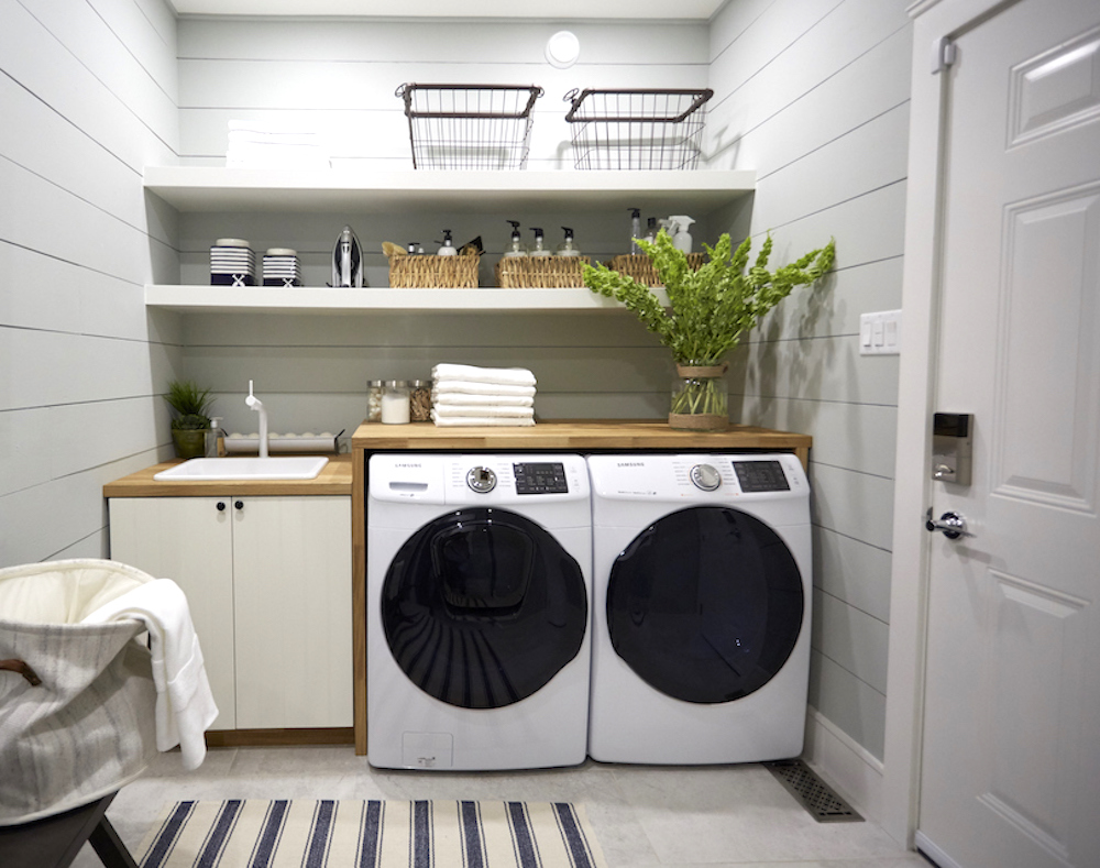 Pretty white laundry room with grey tile floors, a black and white striped rug, a white washing machine and dryer, wooden folding counter and open shelves with laundry soap and accessories as featured on HGTV Canada