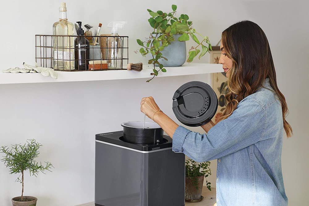 A woman takes the 2-L bucker out a Vitamix FoodCycler set up on a countertop with a shelf above it styled with a basket of kitchen tools and potted plants