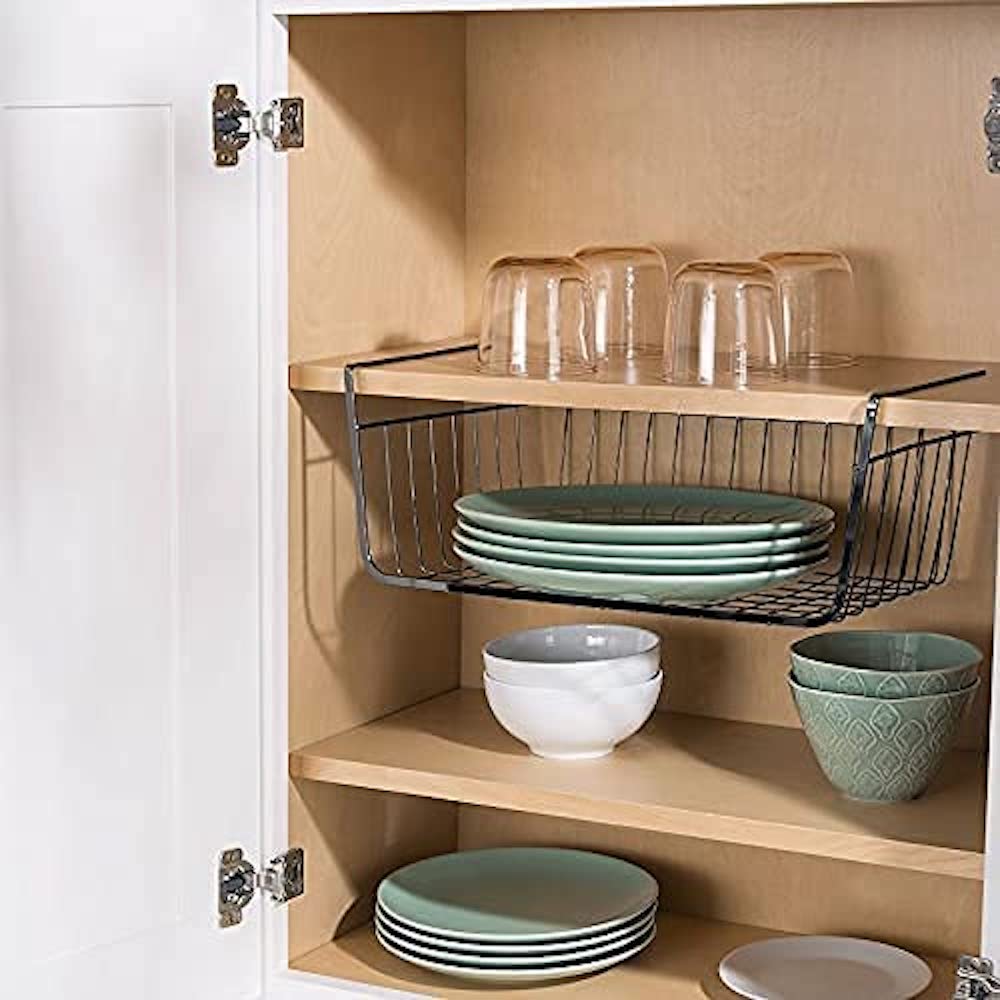 An open kitchen cabinet with a Tebery Under Cabinet Wire Basket hooked onto a shelf with green plates on it and other dishware on the other shelves