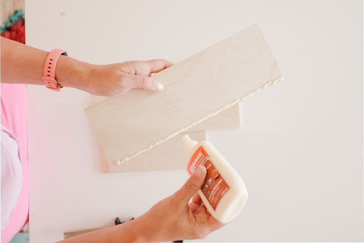 A hand applying wood glue to the bottom of the wide side of a piece of plywood. This piece is one of the two large pieces cut from our original plywood sheet for this DIY. This piece will serve as the DIY organizer's backpiece, the wood glue fixing it to the identical plywood piece used as the organizer's base.