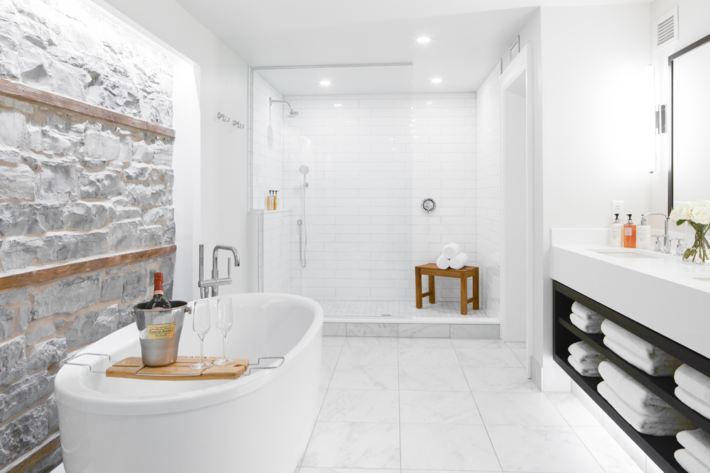 big white bathroom, stone wall on left beside tube with tray of champagne
