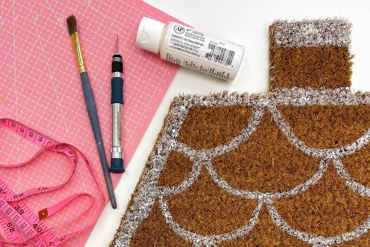 Supplies needed for a gingerbread house doormat: a bottle of white paint, a paintbrush, and an exacto-knife