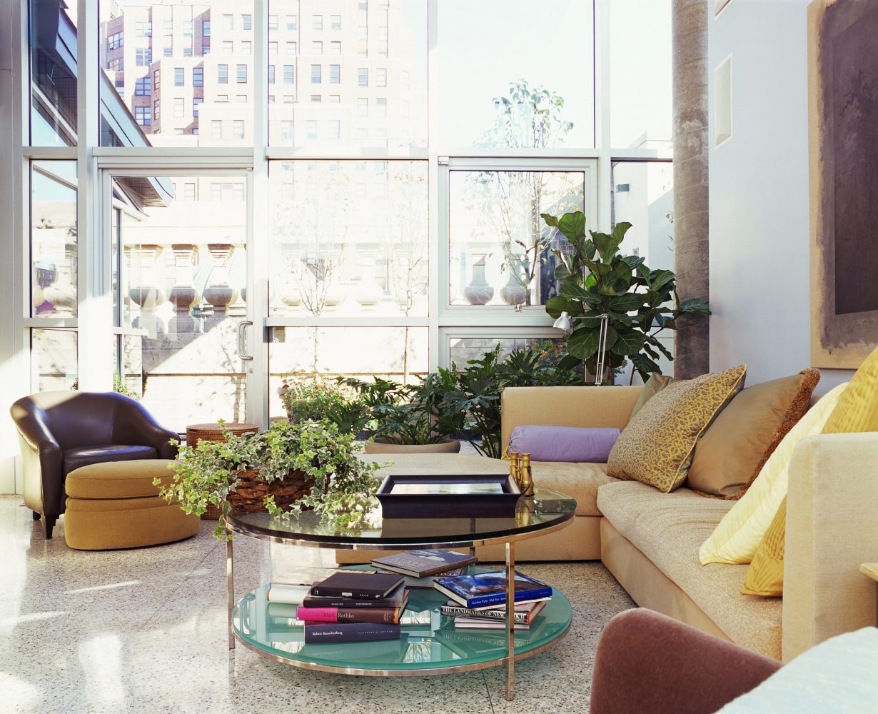 A spacious, modern apartment living room with large glass windows, a glass two-tier table with gold hardware, a buttercream-yellow sectional sofa, some burnt amber armchairs and plenty of leafy green indoor plants.