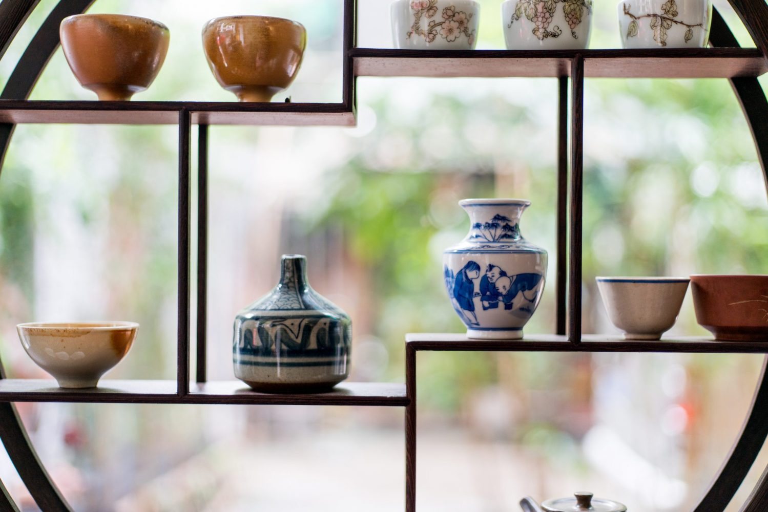 A round, asymmetrical shelf houses several porcelain items, listed in order from top to bottom: A set of three white bowls painted with cherry blossoms; a set of two burnt orange bowls; a pair of mismatched neutral-coloured bowls; a blue and white porcelain urn; a more rustic blue and white vase; and a small beige bowl with neutral-coloured splotches.