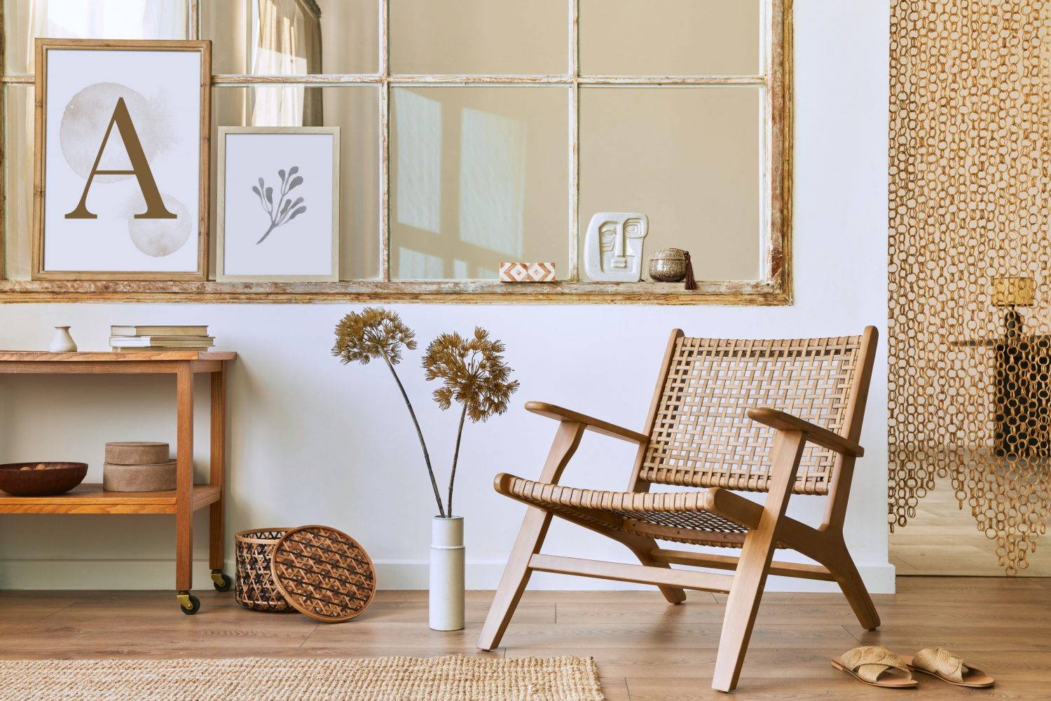 A wood lounge chair with rattan seating and backing is the focal point in this highly neutral room, also featuring wood floors, a woven jute rug, a wicker basket, a two-tier wooden coffee table on wheels with shelves housing decor items, a large French-pane indoor window with rustic trim and some decor elements along the ledge such as a couple photo frames and decorations, and a hanging curtain in the doorway made of chain-linked straw.