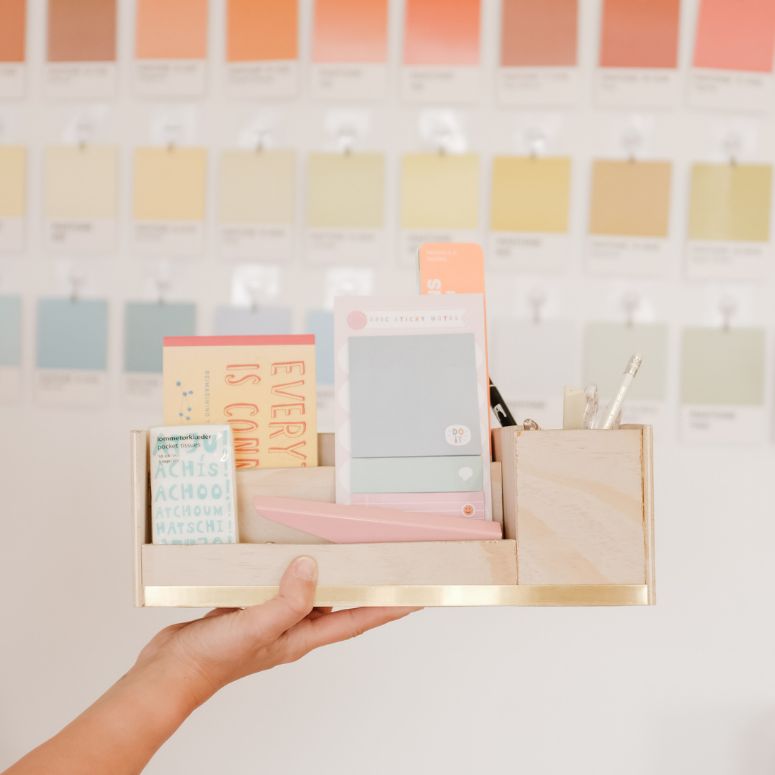 The completed simple wooden DIY desk organizer with a cute copper embellishment at the front, housing pens and notebooks and other stationary items, being held up by a hand in front of a wall decorated with paint swatch cards.