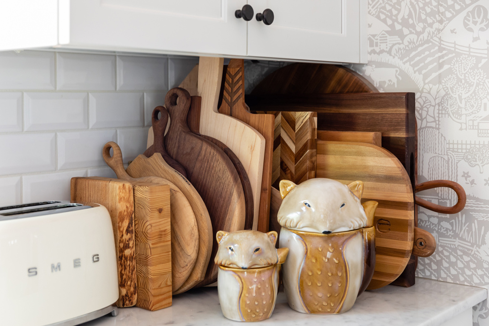 A large collection of wooden cutting boards, two fox-shaped cookie jars and a cream-coloured Smeg toaster in a white kitchen with white subway tile and cream-and-white patterned wallpaper.