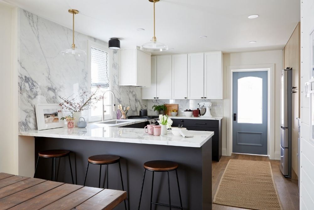 A clean and tidy modern kitchen with a large slab-style marble backsplash over the sink, matching marble countertops, white upper cabinets and black lower cabinets.