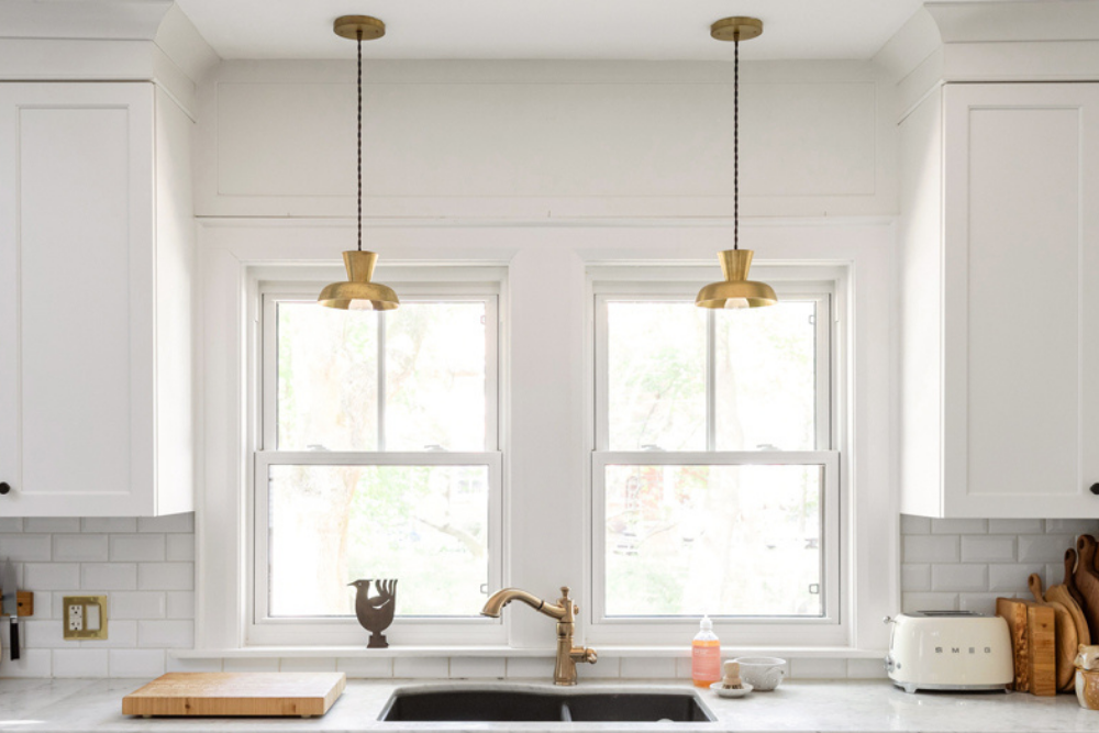 A pair of brass pendant lights hang in front of two twin windows behind the sink in a white kitchen.