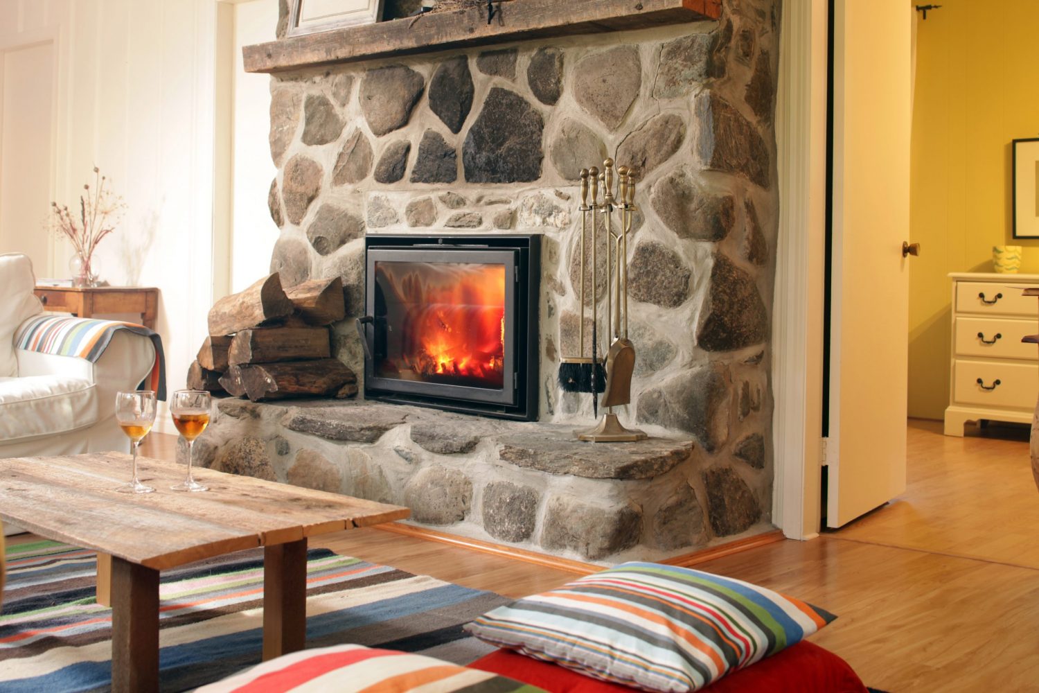 A large stone fireplace with a wide hearth and a roaring fire faces a living room with a white couch, a wooden coffee table and colourful, striped textiles such as a rug, cushions, and a throw over the couch's arm.
